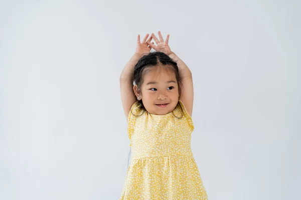 Retrato de preescolar complacido y chica asiática de pie con las manos sobre la cabeza aisladas en gris - foto de stock