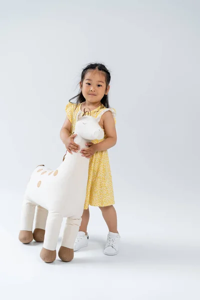 Full length of pleased asian kid holding toy giraffe and standing on grey — Stock Photo