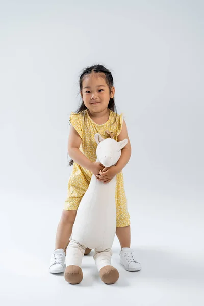 Полная длина счастливого азиатского ребенка, держащего игрушечного жирафа и стоящего на сером — стоковое фото