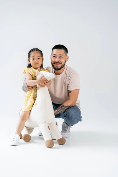 Полная длина счастливого азиатского ребенка, держащего игрушечного жирафа и стоящего рядом с татуированным отцом на сером — стоковое фото