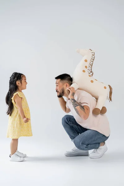 Vista lateral de tatuado asiático homem segurando brinquedo girafa perto de pré-escolar filha no cinza — Fotografia de Stock