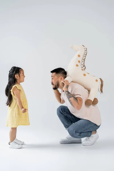 Seitenansicht tätowierter asiatischer Mann mit Spielzeuggiraffe in der Nähe der Vorschultochter auf grau — Stockfoto