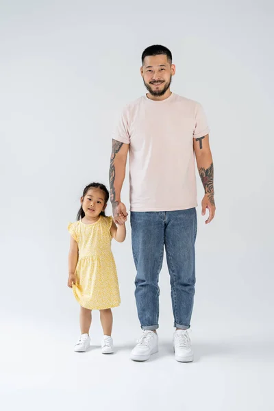 Comprimento total de feliz e tatuado asiático pai segurando as mãos com pré-escolar filha no cinza — Fotografia de Stock