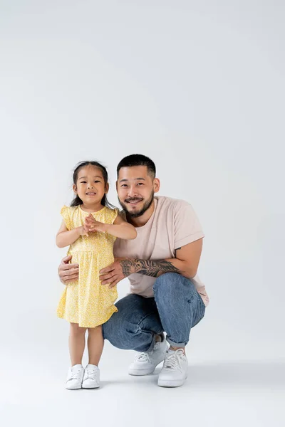 Full length of happy and tattooed asian father hugging preschooler daughter on grey — Stock Photo