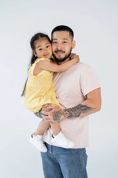 Щасливий і татуйований азіатський батько тримає усміхнену дошкільну дочку ізольовану на сірому — стокове фото