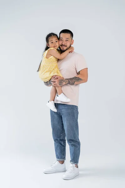 Full length of happy and tattooed asian father holding smiling preschooler daughter on grey — Stock Photo