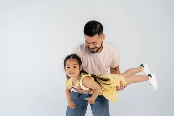Happy asian man holding in arms cheerful daughter in yellow dress isolated on grey — Stock Photo