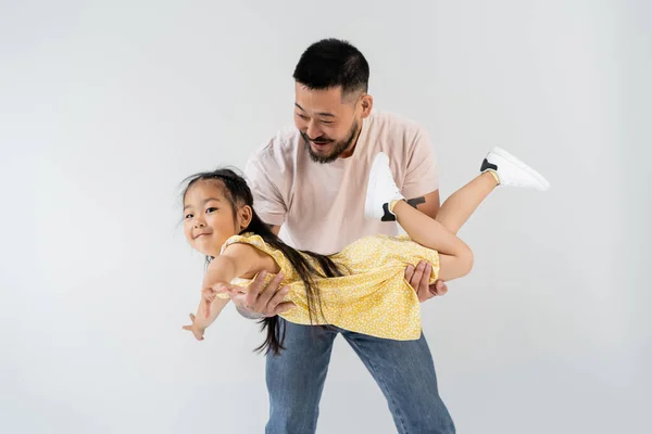 Веселый азиатский мужчина, держащий в руках улыбающуюся дочь в желтом платье, изолированном на сером — стоковое фото