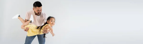 Веселый азиатский мужчина, держащий в руках улыбающуюся дочь в желтом платье, изолированном на сером, баннер — стоковое фото