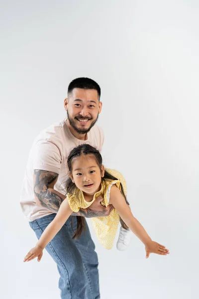 Fröhlicher asiatischer Vater hält freudige Vorschultochter in gelbem Kleid isoliert auf grau in den Armen — Stockfoto