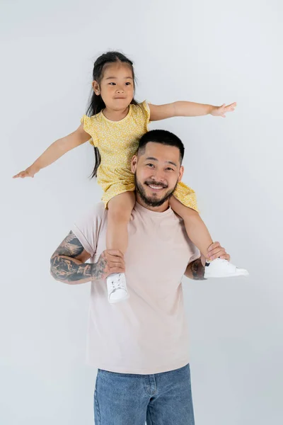 Happy asian child with outstretched hands sitting on shoulders of tattooed father isolated on grey — Stock Photo