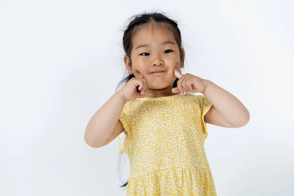 Portrait of preschooler asian girl pointing at cheeks isolated on grey — Stock Photo