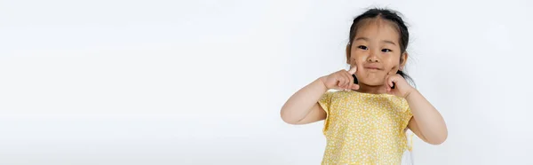 Portrait of preschooler asian girl pointing at cheeks isolated on grey, banner — Stock Photo