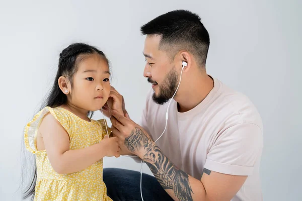 Tatuado asiático hombre usando cableado auriculares cerca hija aislado en gris - foto de stock