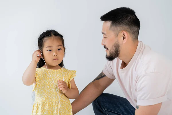 Tattooed asian man looking at preschooler daughter wearing wired earphones isolated on grey — Stock Photo