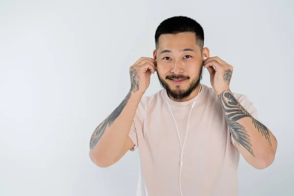 Татуированный азиатский мужчина в футболке, улыбающийся в проводных наушниках и смотрящий на камеру, изолированную на сером — стоковое фото