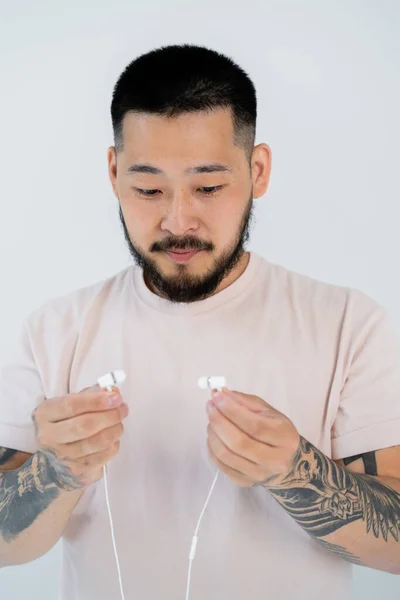 Portrait of tattooed asian man in t-shirt looking at wired earphones isolated on grey — Stock Photo