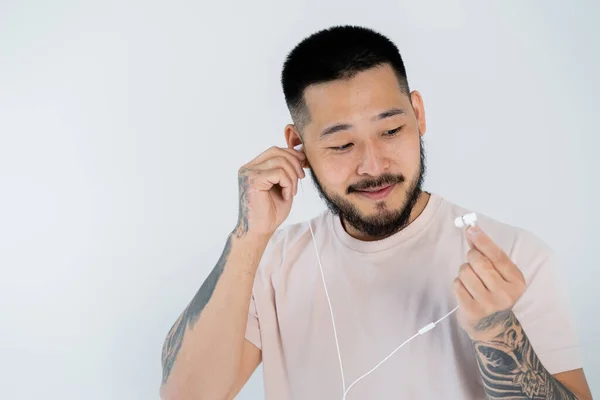Tatuado ásia homem no t-shirt sorrindo e vestindo fios fones de ouvido isolado no cinza — Fotografia de Stock