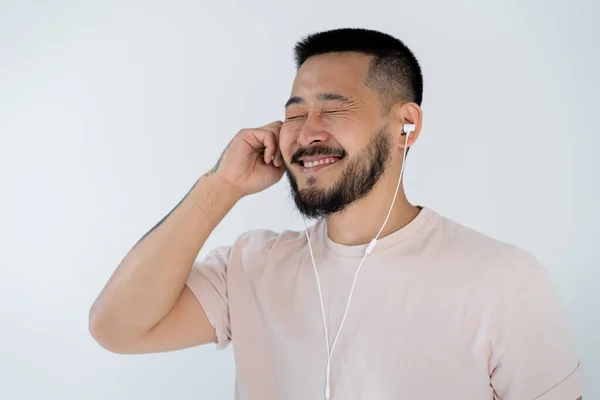 Happy and tattooed asian man with closed eyes wearing wired earphones isolated on grey — Stock Photo