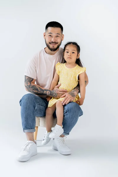 Pleine longueur de heureux asiatique gosse assis sur genou de père en jeans sur gris — Photo de stock