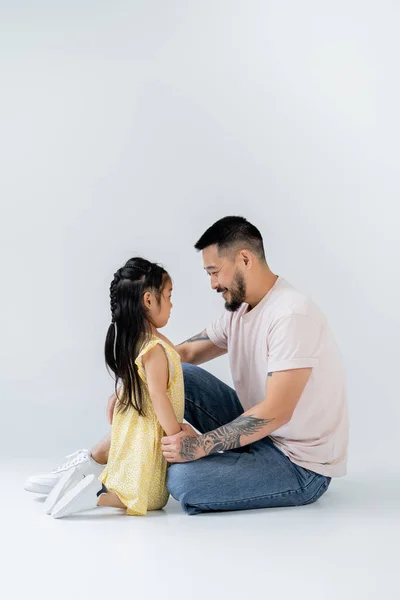 Longitud completa de feliz asiático niño sentado cerca alegre padre en jeans en gris - foto de stock