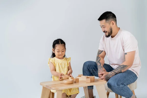 Happy asian man looking at brunette daughter playing with wooden toys isolated on grey — Stock Photo