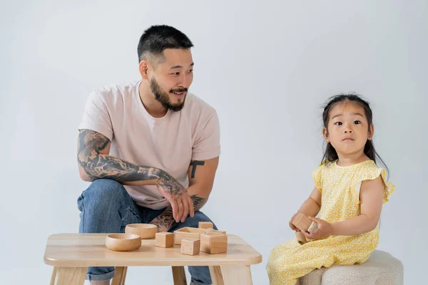 Cheerful and tattooed asian man looking at brunette daughter playing with wooden toys isolated on grey — Stock Photo