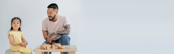 Cheerful and tattooed asian man looking at brunette daughter playing with wooden toys isolated on grey, banner — Stock Photo