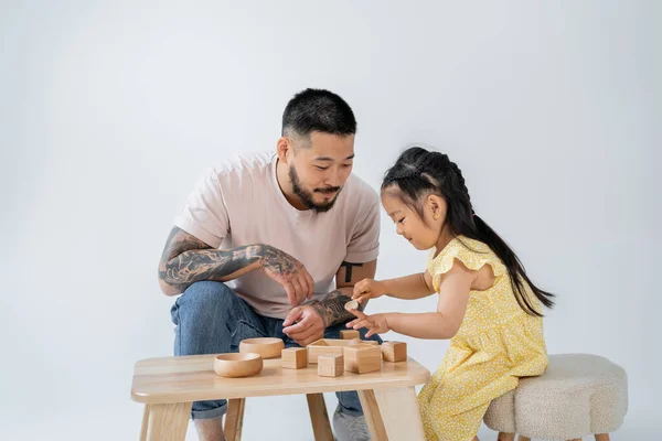 Tattooed asian father playing wooden toys with brunette daughter isolated on grey — Stock Photo