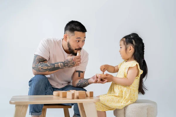 Татуированный отец-азиат играет в деревянные игрушки с дочерью-дошкольницей, изолированной на сером — стоковое фото