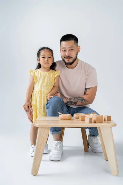 Fröhlicher und tätowierter Vater und asiatische Vorschultochter am Tisch mit Holzspielzeug auf grau — Stockfoto
