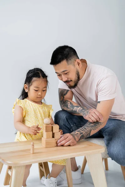 Татуированный отец-азиат играет в деревянные кубики с дочерью-дошкольницей, изолированной на сером — стоковое фото