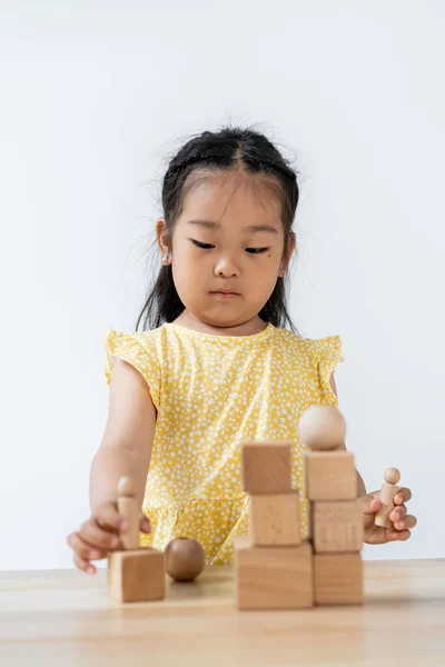 Niño preescolar asiático en vestido amarillo jugando con formas de madera borrosas aisladas en gris - foto de stock