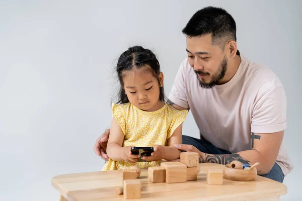 Tattooed asian man looking at brunette daughter using smartphone near wooden toys isolated on grey — Stock Photo