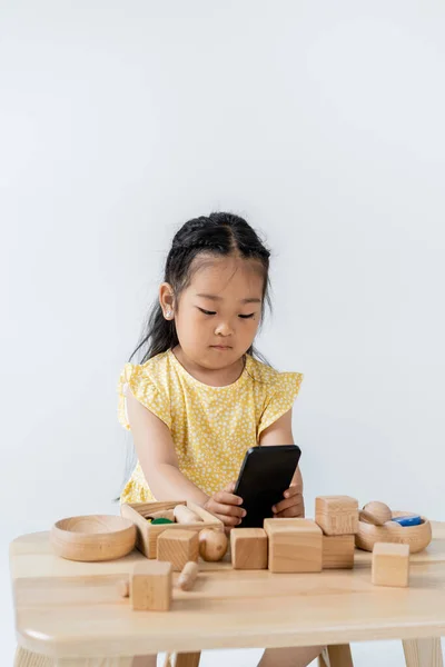 Asiático chica usando smartphone cerca de madera juguetes en mesa aislado en gris - foto de stock