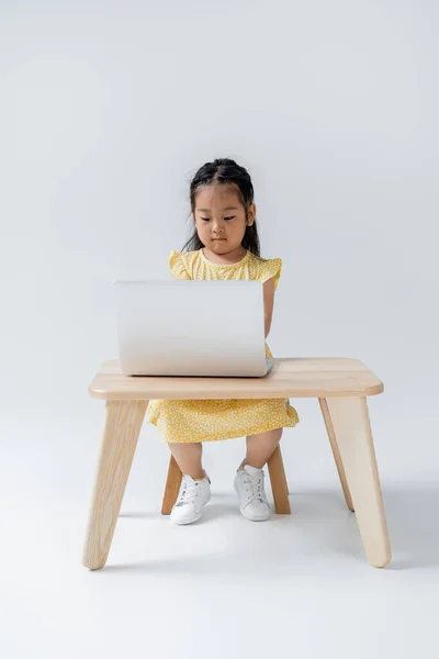 Pleine longueur de fille asiatique en robe jaune en utilisant un ordinateur portable tout en étant assis à la table en bois sur gris — Photo de stock