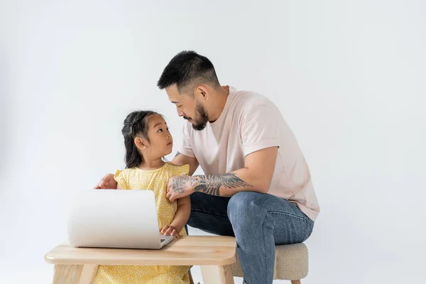 Tätowierter Vater blickt überraschte asiatische Tochter in der Nähe von Laptop auf Holztisch isoliert auf grau — Stockfoto