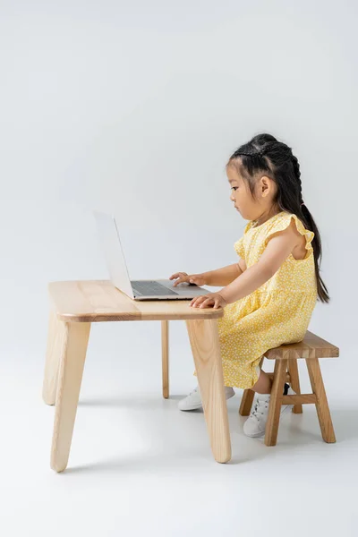 Vue latérale de fille asiatique en robe jaune à l'aide d'un ordinateur portable sur gris — Photo de stock
