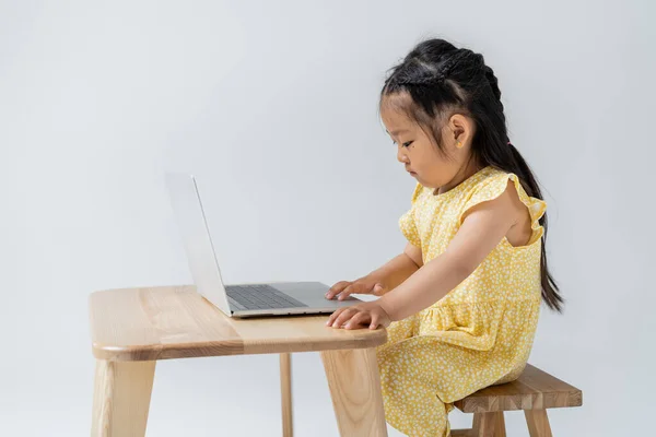 Vue latérale de la fille maternelle asiatique en robe jaune en utilisant un ordinateur portable isolé sur gris — Photo de stock