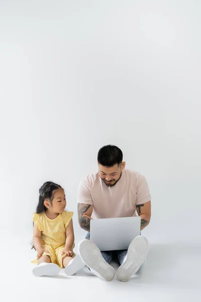 Feliz asiático padre trabajando remotamente en portátil cerca hija en gris - foto de stock