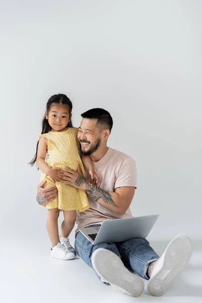 Alegre asiático pai sentado com laptop perto filha no cinza — Fotografia de Stock