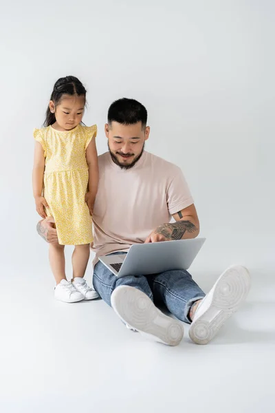 Alegre asiático pai usando laptop perto filha no amarelo vestido no cinza — Fotografia de Stock