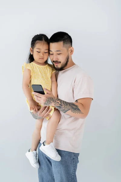 Asiatischer tätowierter Mann benutzt Smartphone und hält Tochter im Arm isoliert auf grau — Stockfoto