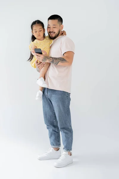 Повна довжина щасливого татуйованого чоловіка за допомогою смартфона і тримає азіатську дочку в руках на сірому — стокове фото