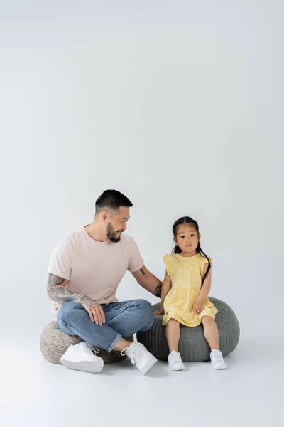 In voller Länge glückliche tätowierte Vater und asiatische Tochter in gelbem Kleid sitzen auf Puffstühlen auf grau — Stockfoto