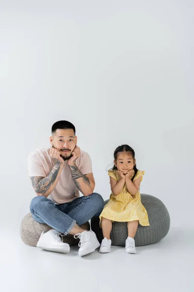 Full length of tattooed father and asian daughter in yellow dress sitting on puff chairs on grey — Stock Photo