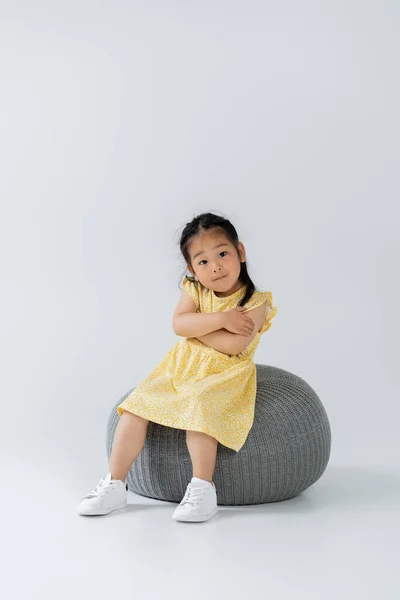 Full length of asian kid in yellow dress sitting on puff chair on grey — Stock Photo