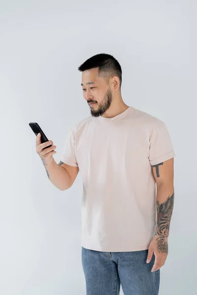 Smiling asian man with tattoos using smartphone isolated on grey — Stock Photo