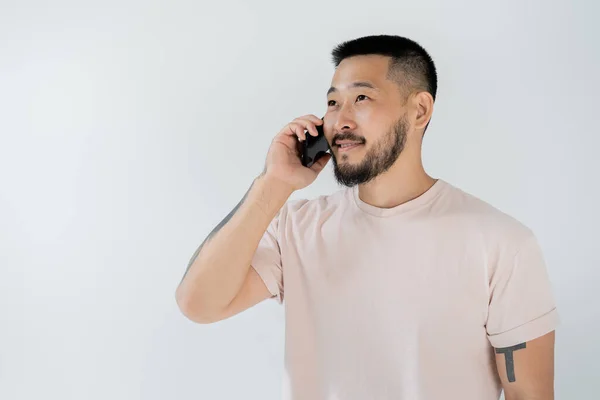 Усміхнений азіатський чоловік з татуюваннями говорить на смартфоні ізольовано на сірому — стокове фото