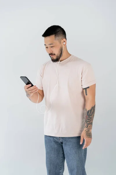 Tattooed asian man in wired earphones listening music and holding smartphone isolated on grey — Stock Photo